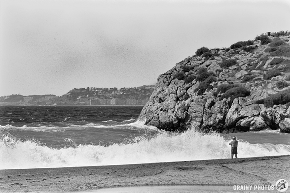 Black and white photo of Salobreña beach shows waves crashing against the rocky coastline; a person stands near the water watching the waves, with distant hills and buildings visible in the background.