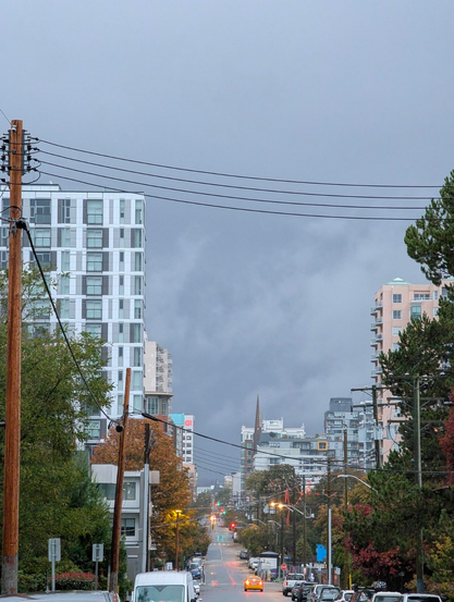 A shot down view street. Looking west towards downtown, and an ominous rain wall behind it.
The street is lined with trees that are in the middle of changing colour, and high density (apartment/Condon/office) buildings.
Early morning. Street lights are still on.