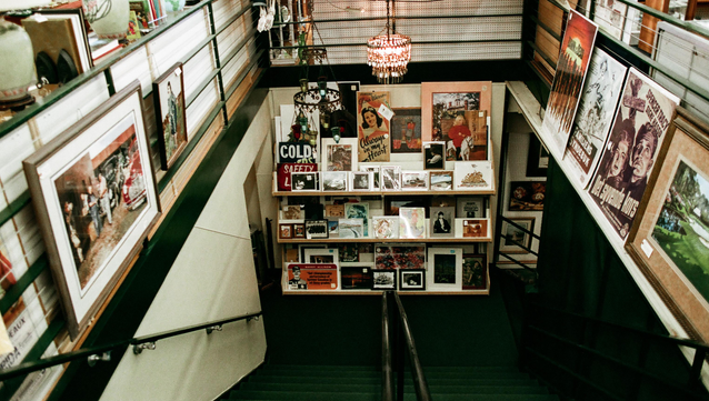 A photo taken inside a two story antique shop.  The shot is taken from the top of two way stairs leading to a lower level.  The inside of the stairwell has framed artwork on both walls and a rack holding flat items such as playbills, photographs and unframed artwork at the bottom.