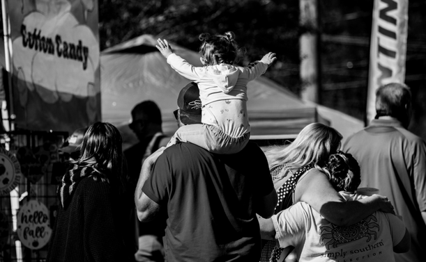 This is a black and white photo of a group of people looking away from the camera.  There is a little girl sitting on her father's shoulder with both of her arms raised into the air.  Just to their right are two people giving each other a tight one arm hug.