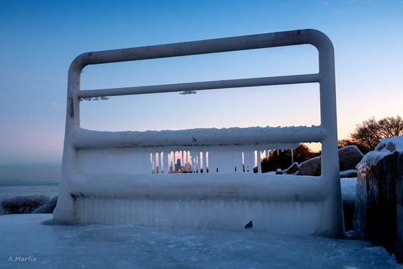 A color photograph on the Chicago lakefront near Montrose Harbor. Pictured is a frozen guard rail and beyond is Lake Michigan and the Chicago skyline. It is sunset and the sky is blue and purple. Circa February 2020.