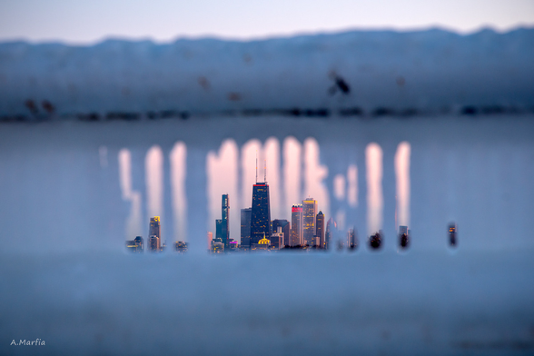 A color photograph on the Chicago lakefront near Montrose Harbor. Pictured is a close up of a frozen guard rail which you can see through to the Chicago skyline. It is sunset and the sky is blue and purple. Circa February 2020.