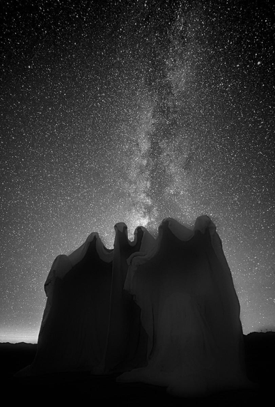 A black and white portrait photo of the Milky Way galaxy extending out of the heads of an outdoor sculpture of three ghostly figures in capes. The Milky Way extends straight up and is a very dense field of tiny dots of stars. 
