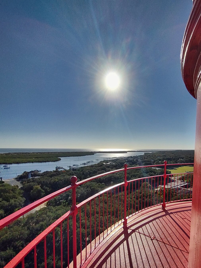 Sun shines brightly over a scenic coastal landscape viewed from a red lighthouse balcony. A river and lush greenery extend toward the horizon.