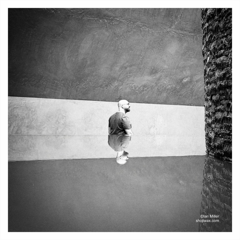 Black and white photo of the upper half of a man reflected in a pool of water as he walks down a ramp.