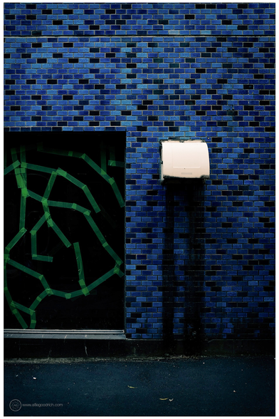 An alleyway in Higashi-ginza, Tokyo. The walls are tiled with small tiles of various blues from light to deep indigo. An extractor fan is mounted on the wall. From it dark streaks of dirt run down to the ground. An indent in the wall. to its left, is painted black and covered with a pattern of yellow tape. 