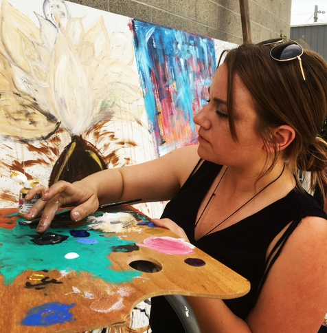 A Hueman actively painting with brown hair in a bun, wearing a black tank top and sunglasses on my head. I’m dipping my fingers in paint on a palette. A mural shared with friends is in the background