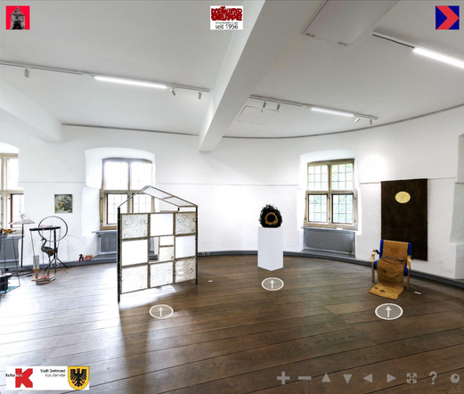 A room with curved, white walls in which medieval windows are set. Green foliage can be seen through the windows. The room has a wooden floor made of thick dark brown planks. There are sculptures a hut standing in the room and paintings hanging on the walls. 