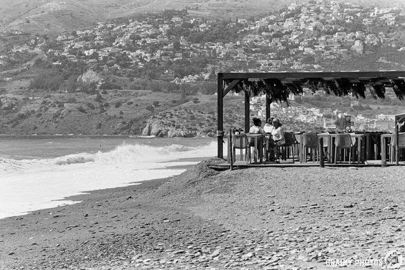 A black and white photo of people sitting in a shaded chiringuito on Salobreña beach, overlooking waves, with the hillside town of Salobreña visible in the background.