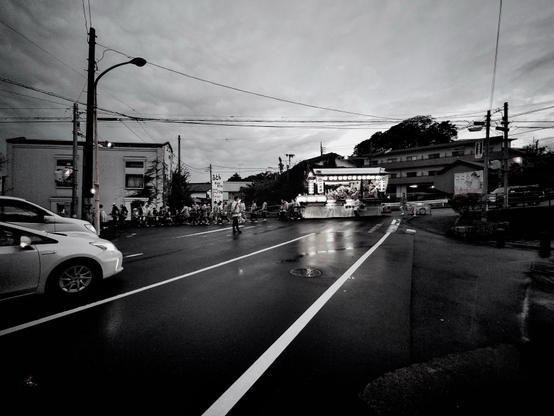 To the left, two cars are being held up by a man wearing a hapi jacket (waist length light cotton festival attire.) Behind him is a float with a roof, rows of lanterns and a band of drummers and flautists. The float is being maneuvered down hill at a road junction by a group of about twenty people also in hapis. White road markings draw the eye towards the festive group. It has recently rained so reflections from the lanterns play across the wet tarmac. The leaden sky is grey and heavily clouded. Telephone lines, street lamps and residential buildings populate the background.