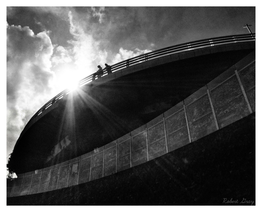 Two distant figures look down from a curved railing. They are backlit by the sun and therefore seen in silhouette.