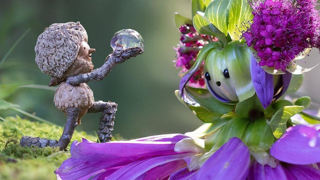 A Becorn figure made from acorns and twigs kneels on moss while presenting a water droplet to a flower character constructed from purple dahlia petals and green leaves.