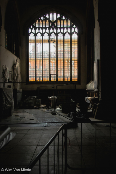 Church interior.  The lights are off so it is tenebrous inside, drawing attention to the gothic window; the orange is from the brick houses opposite.  Restoration work is going so so parts of the nave are fenced off.