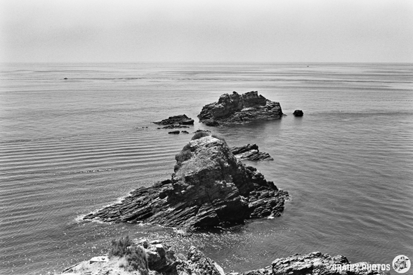 Black and white photo of rocky outcrops near Salobreña, extending into a calm sea, with gentle waves and a hazy sky in the background. The coastline scene feels serene and timeless.