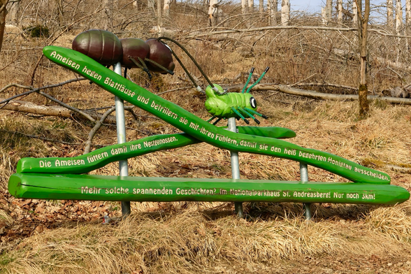 Am Wegesrand im Wald steht ein übergroßes Model einer Ameise auf einem grünen Ast. Vor der Ameise ist eine übergroße Blattlaus auch auf dem grünen Ast. Insgesamt sind drei Äste zu sehen. Auf diesen ist in kurzen Sätzen erklärt, warum Ameisen die Blattläuse pflegen. 