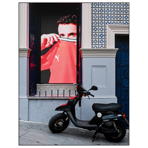 The image shows an urban street scene. A black scooter is parked on the sidewalk in front of a building. The building's facade features a large screen in a window or doorway, showing a video of a person wearing the red sweatshirt of the portuguese national football team. The facade of the building is decorated with blue and white patterned tiles, typical of Portuguese azulejos, around the door and window frames. The door is painted blue with decorative ironwork. The overall scene is set in daylight. (draft by mistral.ai, revised by author)