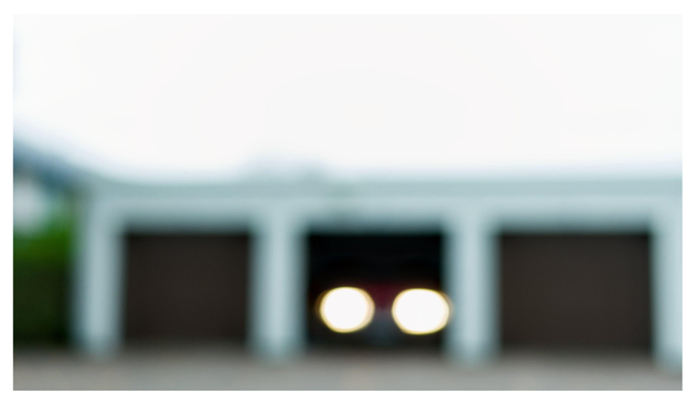 En liggande bild på en röd bil på väg att lämna ett garage. De två garagen på båda sidorna är stängda. Till vänster anas en huslänga med gröna växter. Allt är ordentligt ur fokus.