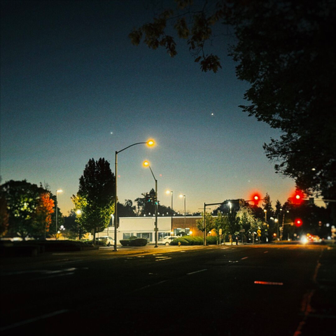 Street lamps light up a city street under a twilight pre-sunrise, as stars and planets dot the sky.