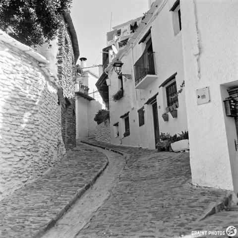A narrow cobblestone street lined with white-washed buildings, potted plants, and balconies curves uphill in Capileira, Spain, a charming Mediterranean village under a bright sky.