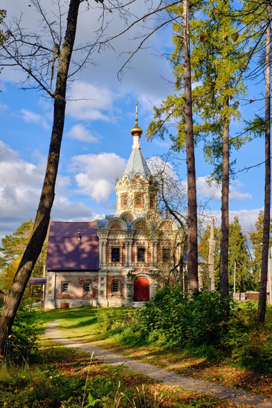 A historic church with ornate architecture, featuring a blue and gold dome, is surrounded by trees and greenery. A path leads to the entrance, under a partly cloudy sky.