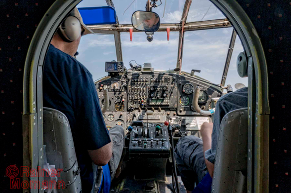 Farbfotografie im Format 3:2 Landscape.
Blick in das Cockpit einer Antonov AN2 und weiter, durch die Scheiben in den blauen, mit wenigen Schleierwolken bedeckten Himmel. Im Cockpit sind viele Knöpfe, Schalter und Hebel zu sehen. Mit dem Rücken zur Kamera und größten teils durch die Trennwand zum Cockpit abgedeckt, links der Pilot mit Baskape und Kopfhörern und rechts der Copilot, ebenfalls mit Kopfhörer.