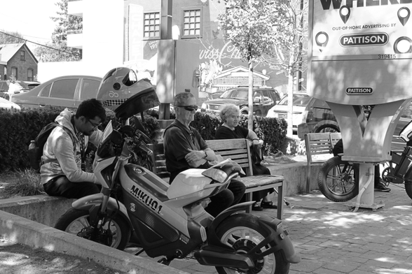 In a small sitting area of outdoor benches, three people are visible. A young man sits by his motorbike, looking down at his phone. An older man with very dark glasses sits ramrod straight, looking ahead fixedly. An older woman looks toward the camera, realizing she’s being photographed. Embedded in the paving tiles is a huge advertising sign with just the bottom of the slogan showing. 
