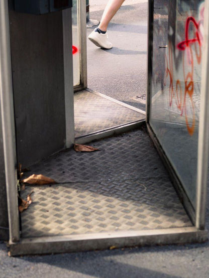 A leg of a man walking is visible through a metal and glass phone booth.