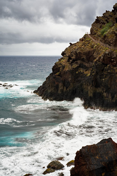 Fotografía de un paisaje oceánico, en la parte superior central derecha una masa de tierra volcanica se recorta contra el cielo y el oceano, una barandilla revela el camino por el que se accede subiendo por uno de sus costados. En la esquina inferior derecha una roca volcanica con colorido rojo negro y naranja aparece en primer plano. La mayor parte del lado izquiero del cuadro la ocupa el oceano, con colores turquesas azules y morados, asi como el blanco de la espuma de las olas al romper contra las rocas. La linea del horizonte en el tercio superior separa el oceano de las nubes grises del cielo encapotado.