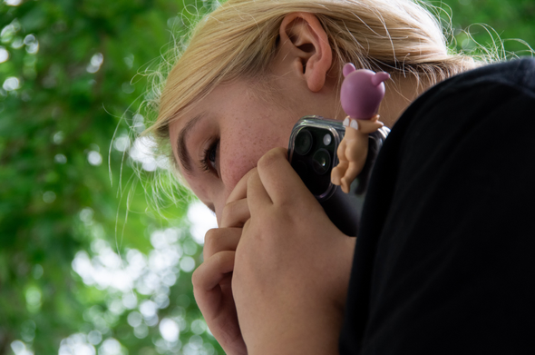 A photo of a young lady shot from her lower left.  She is holding an IPhone with a cherub attached in her hands which partially covers her face.