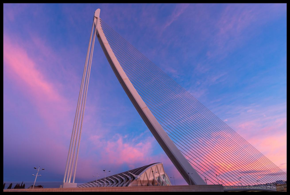 The modern cable-stayed Pont de l'Assut de l'Or bridge near Valencia's City of Arts and Sciences photographed at sunrise, showing its distinctive white asymmetric pylon and radiating cable stays against a spectacular sky painted in shades of pink, purple, and blue.