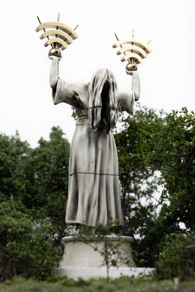 A ghostly statue with long hair, draped in a flowing robe, raises two golden Wi-Fi symbol sculptures against a cloudy sky.