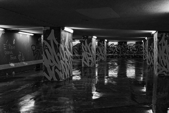 Fotografia a preto-e-branco de uma decadente passagem subterrânea. Os pilares e as paredes estão cobertos por graffiti, podendo ler-se, na parede à esquerda, a frase: «Tudo é uma fase.» O chão molhado reflecte a luz das lâmpadas fluorescentes, pejadas de teias de aranha.