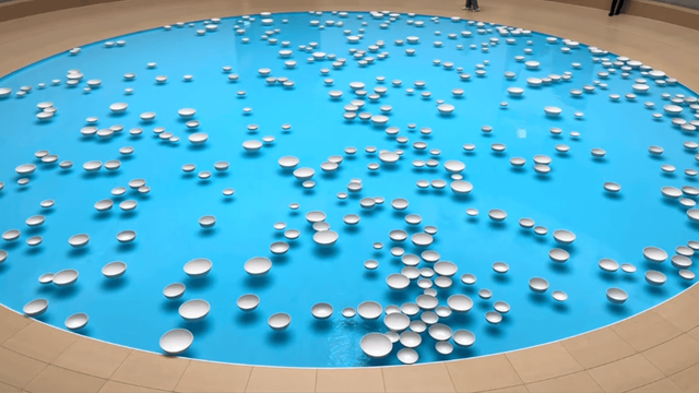 White porcelain bowls float across a circular pool of bright blue water in an indoor installation space with beige flooring.