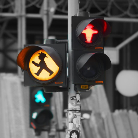 Eine rote Fußgängerampel im Vordergrund, mit einem gelben Warnlicht für abbiegende Autos. Dahinter unscharf eine grüne Fußgängerampel und monochrome Gerüste.