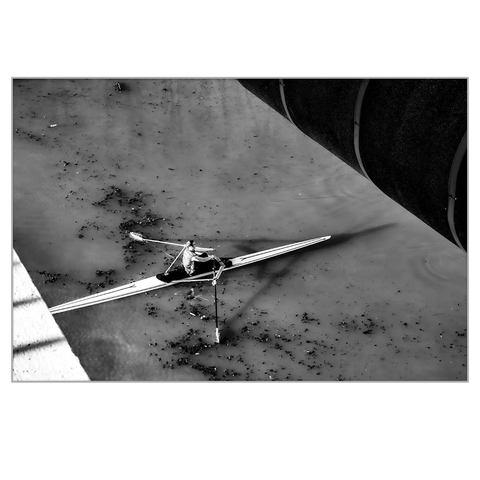 A white kayak photographed from above. A white section of the bridge is visible in the lower left corner of the image, and a black section is visible in the upper right corner, framing the black-and-white image. The kayak is moving diagonally.