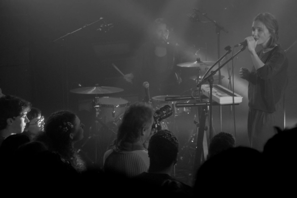 Monochrome, black-and-white photograph of a female singer on stage in front of the crowd, including a photograph with a large lens and three young people with their faces in the stage lights. In the background, a drum kit is being played by a woman who is also singing backup vocals.
