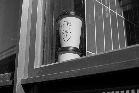Black-and-white photo of a coffee cup with, "Coffee here!" written on it along with a smiley face. The coffee cup has been placed on a cafe window sill.