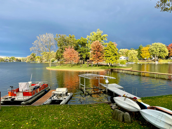 Photography of a small island on a suburban lake with boats and docks and a lawn in the foreground. The island is well lit with some trees having turned orange. There are dark clouds in the background.
