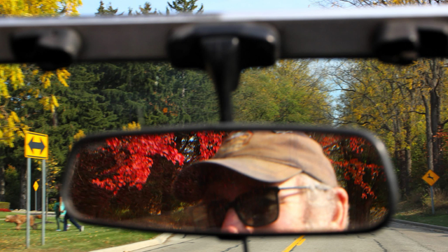 Photograph of a man with a baseball cap and sunglasses in a rearview mirror with red leaves behind him to the front of the car is a road with green and yellow trees.