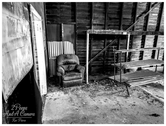 A black and white photograph of a dark, worn armchair sitting alone on a dusty concrete floor inside a decaying, abandoned wooden shed or workshop.

To the right is a rusty metal shelving unit. An open doorway on the left provides a bright contrast. The image is signed Kev Peirce.