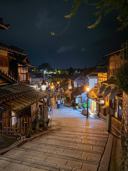 Típica vista de una callejuela del barrio antiguo. El suelo está empedrado. Va bajando la colina rodeada de casas tradicionales de madera. Era de noche con iluminación naranja de las farolas. La calle está casi desierta a estas horas.