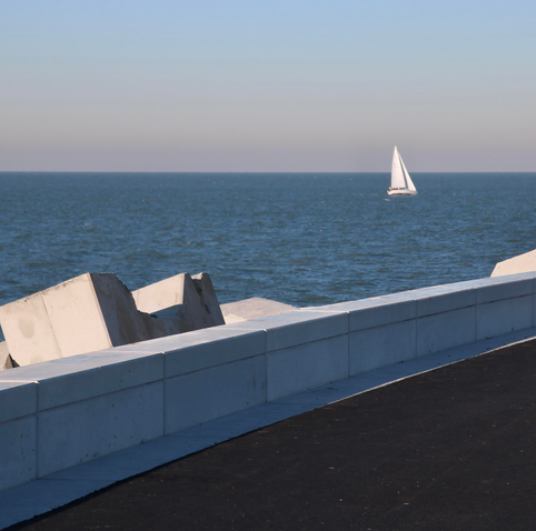 das bild zeigt einen blick auf das meer mit einem segelboot in der ferne. im vordergrund befindet sich eine befestigung, möglicherweise eine promenade oder ein damm, mit weißen betonblöcken. der untergrund ist dunkel, wahrscheinlich asphalt. der himmel ist hell und klar, was auf einen sonnigen tag hindeutet.