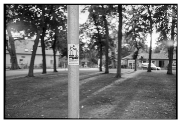 Black and white photograph in landscape orientation. An out of focus park scene fills most of the image. Only a thin pole is in focus. On this pole aa anti wind-energy sticker reading "Keine Windräder im Naturpark Dahme-Heideseen".