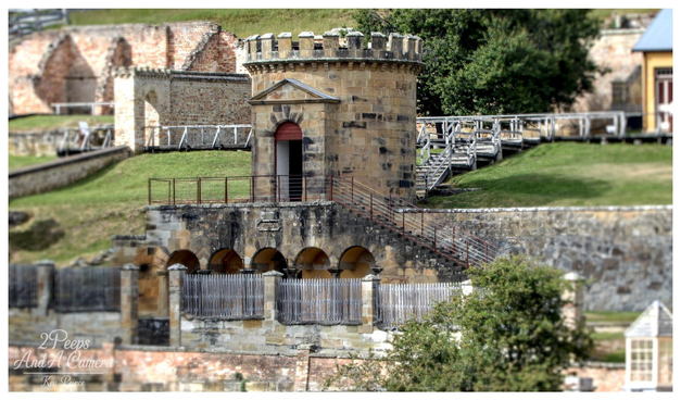 A close up photograph of the sandstone Guard Tower, a small, cylindrical, crenellated building, standing on a fortified stone base with archways at the Port Arthur historic site in Tasmania.

A steep wooden staircase leads up to the tower. The surrounding grass is green, and the ruins of larger brick buildings are visible in the blurred background.