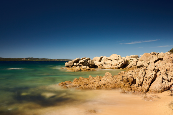 The photo shows upfront a sandy beach with rock formations coming from the right into the sea. The rocks are in a warm amber tone. The blue greenish water around seems like a foggy plane due to the long exposure. The is dark blue except from two light strokes of white lines of clouds far away to upper right..