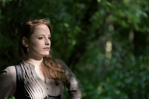 A young woman with red hair hanging on her left shoulder, with her hands on her hips out of frame, looking far away to the right of the pic. The background is filled with trees’ leaves