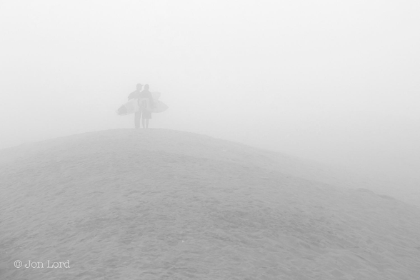 This is a black and white photo in landscape format of two surfers carrying their surfboards and standing on a sand dune in thick fog. Hermosa Beach, California (2014).
Rising up from the base of the image towards the middle is the crest of a sand dune that falls away slightly to the left and more so to the right. The view is obscured by a thick sea fog reducing the visibility to a hundred metres or probably less. Standing on the crest of the dune and to the left and slightly above centre are two morning surfers standing together. Both are carrying their surfboards and are looking towards the sea. Above the dune and surfers is a sky totally obscured by the fog. 
The dune is not natural but a berm, dyke or levee that has been bulldozed to stop winter storms causing the waves to run up the beach and into the town and beach-front houses.