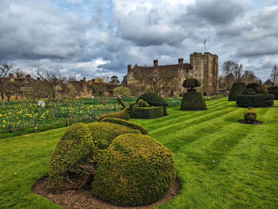 A beautifully manicured garden features an array of imaginative topiary sculptures, including abstract shapes and animal forms, set against a backdrop of lush green lawns. Vibrant yellow daffodils bloom in organised rows, adding a splash of colour to the scene. In the distance, a historic stone castle with a flag flying atop its tower stands majestically, surrounded by traditional stone buildings with steep roofs and chimneys. The sky overhead is filled with dramatic, moody clouds, suggesting an overcast day. The garden is expansive, with neatly trimmed hedges and pathways leading the eye towards the castle, creating a harmonious blend of nature and architecture.