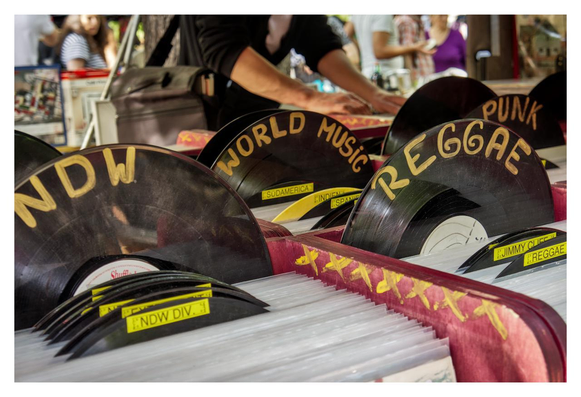 A close-up photo of a box filled with vinyl records at a flea market. In the foreground, several black vinyl record dividers are prominently visible, each labeled in handwritten-style yellow text. From left to right, the visible labels read "NDW" (standing for Neue Deutsche Welle, a German music genre of the 80's), "WORLD MUSIC", "REGGAE", and "PUNK". The records themselves are black LPs in white or clear sleeves, stacked upright in a red wooden box decorated with yellow star-like symbols. In the background a person's arm and hand, wearing a black long-sleeve shirt, are reaching into one of the boxes, flipping through the records.