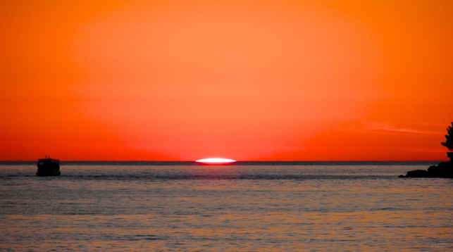 Dieses Foto zeigt einen Sonnenuntergang über dem Meer. Der Himmel ist in intensiven Rottönen gefärbt, während die Sonne tief am Horizont steht und ihr Licht auf das Wasser wirft. Im Vordergrund sieht man das ruhige Meer, das die Farben des Himmels widerspiegelt. Auf der linken Seite des Bildes ist eine kleine, dunkle Silhouette - ein Boot - zu sehen. Rechts im Bild erkennt man die Umrisse von Felsen einer Landzunge. Die Stimmung des Bildes ist friedlich.